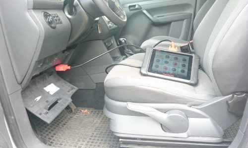 Interior view of a vehicle with a diagnostic device on the car seat, which is connected to the vehicle via a cable. The cover of the fuse box has been removed.