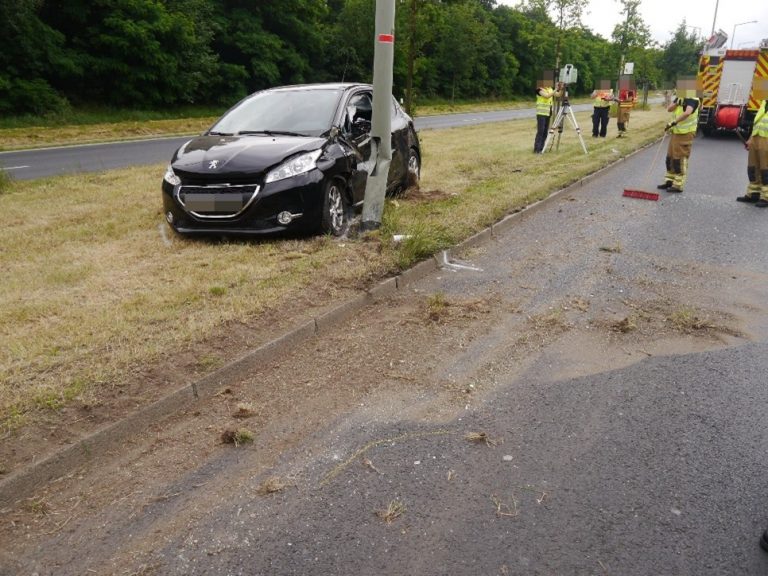 Traffic accident involving a black car that has crashed into a lamppost while emergency services investigate the scene on a nearby road.