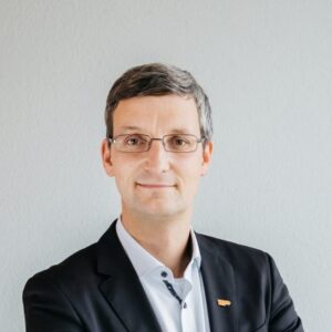 Portrait of Henrik Liers, Managing Director of VuFo, in a dark suit with a white shirt and glasses, against a light-coloured background.