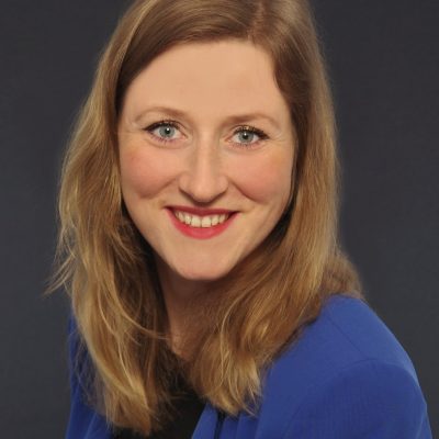 Portrait photo of Susanne Lindner, smiling and looking into the camera. She has shoulder-length blonde hair and is wearing a blue top against a dark grey background.