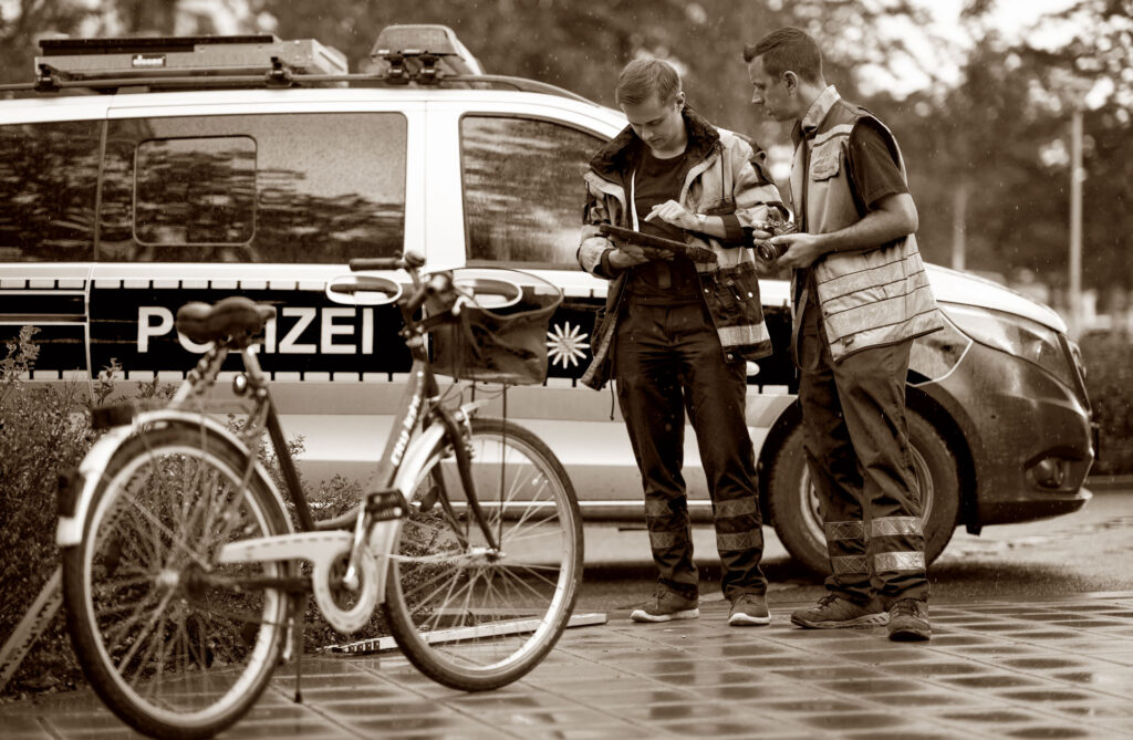 Zwei Mitarbeiter der VuFo stehen vor einem Polizeifahrzeug und betrachten gemeinsam ein Dokument. Im Vordergrund sind zwei Fahrräder zu sehen. Die Szene ist in Sepia gehalten und zeigt eine typische Situation am Unfallort.