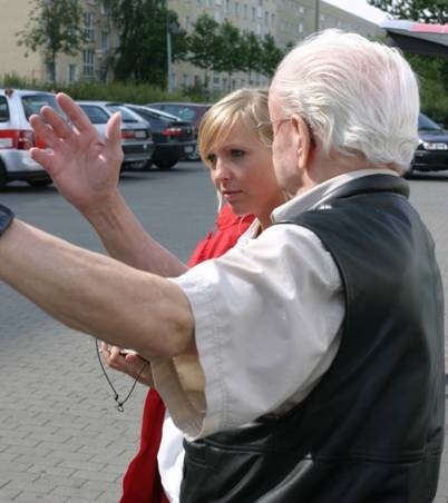 Zwei Personen, eine Frau und ein älterer Mann, die sich unterhalten und gestikulieren, im Freien auf einem Parkplatz.