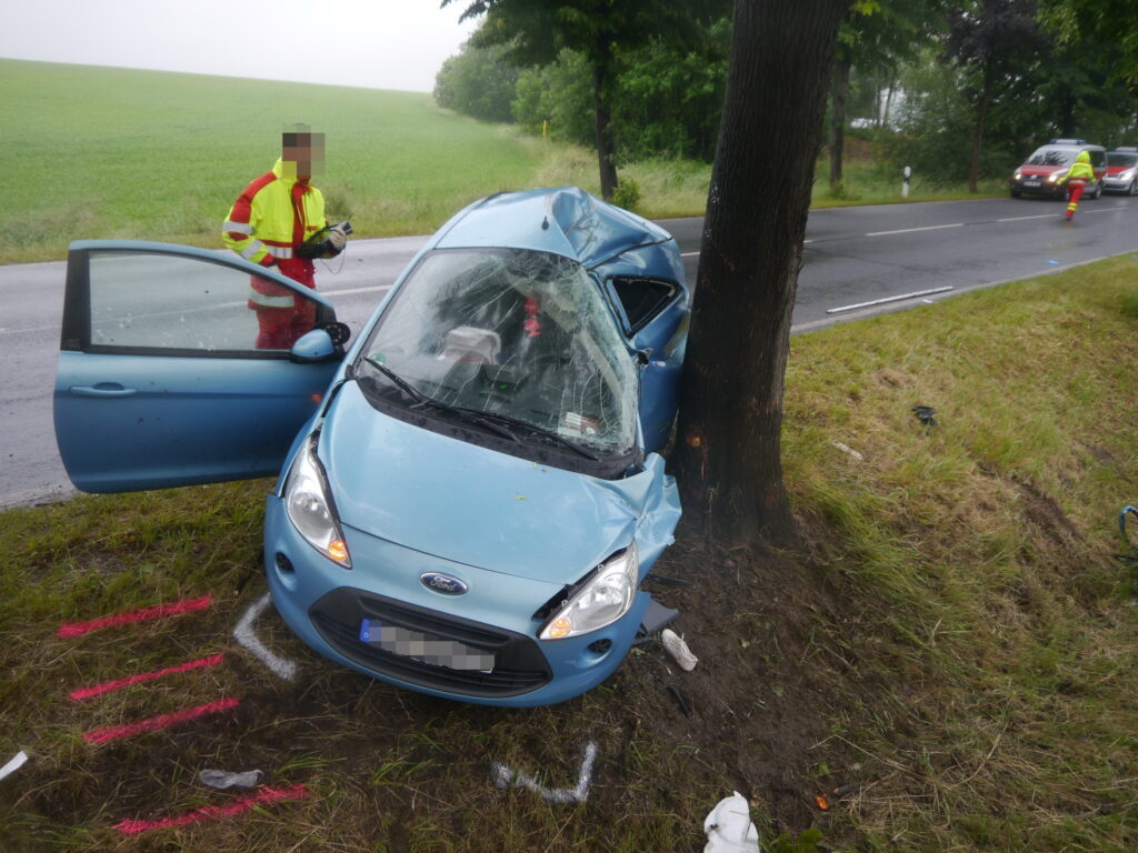 Traffic accident involving a blue car that has crashed head-on into a tree, with a paramedic in the background on a country road.